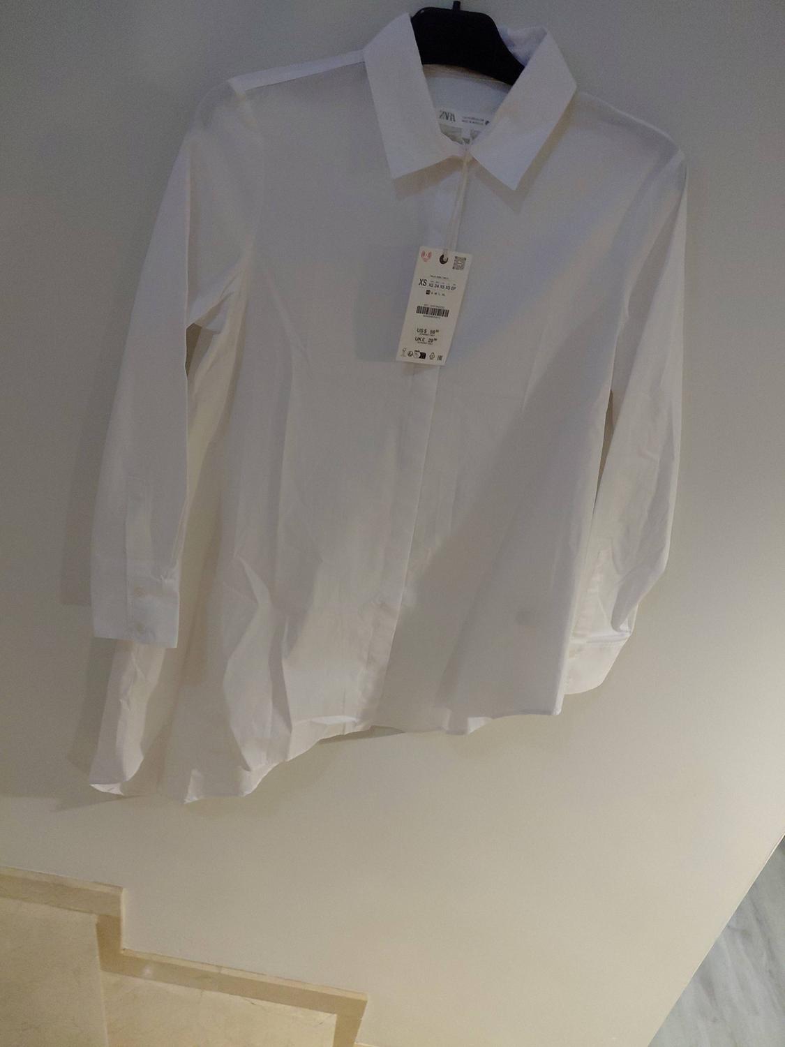 White long-sleeved shirt with an asymmetric hem hanging on a hanger. Label with the text “ZARA”, “XS”, “US $39.90”, “UK £29.99”.