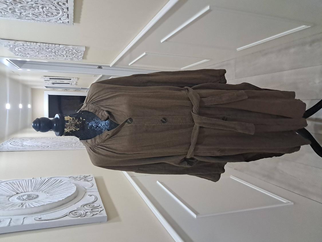 Mannequin in a light brown fabric coat with a belt, in a hallway with white panelled decor on the walls.