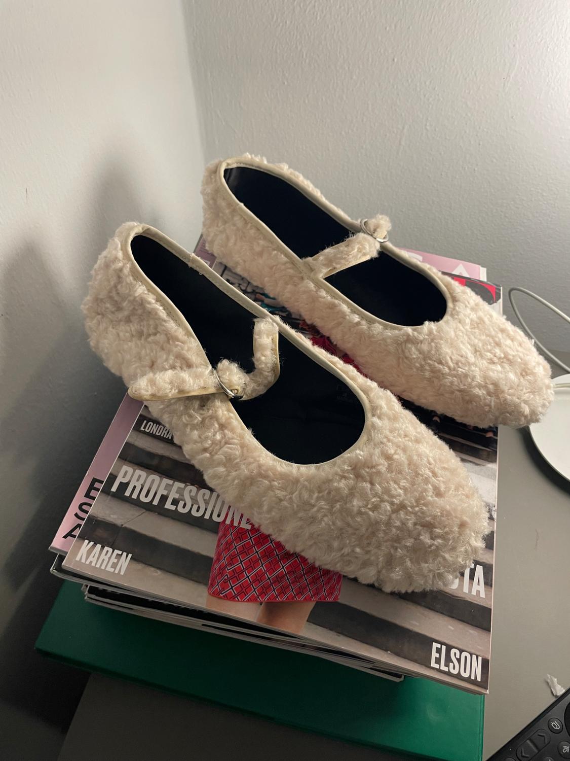 Beige fluffy slippers with a strap on top of stacked magazines on a grey table. Visible text: "PROFESSIONISTA", "KAREN", "ELSON".