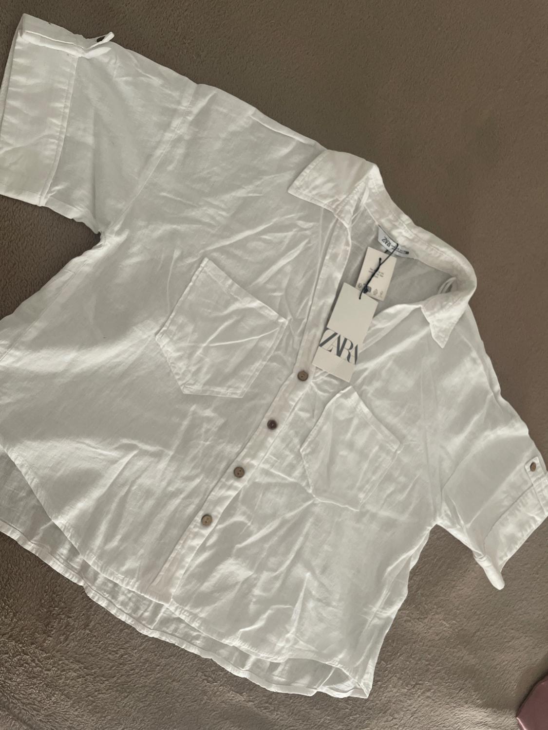 Short-sleeved white shirt with buttons and two pockets, laid out on a brown background. Includes a label with the text ZARA.