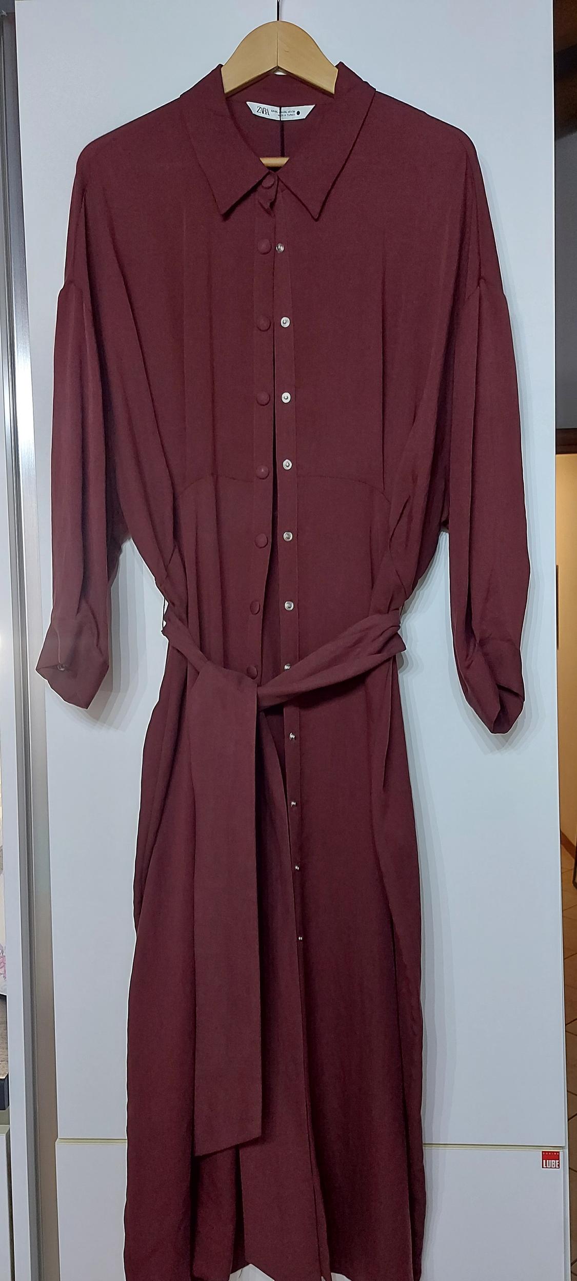 Burgundy long shirt dress with front buttons, adjustable belt and long sleeves, hanging on a wooden hanger.