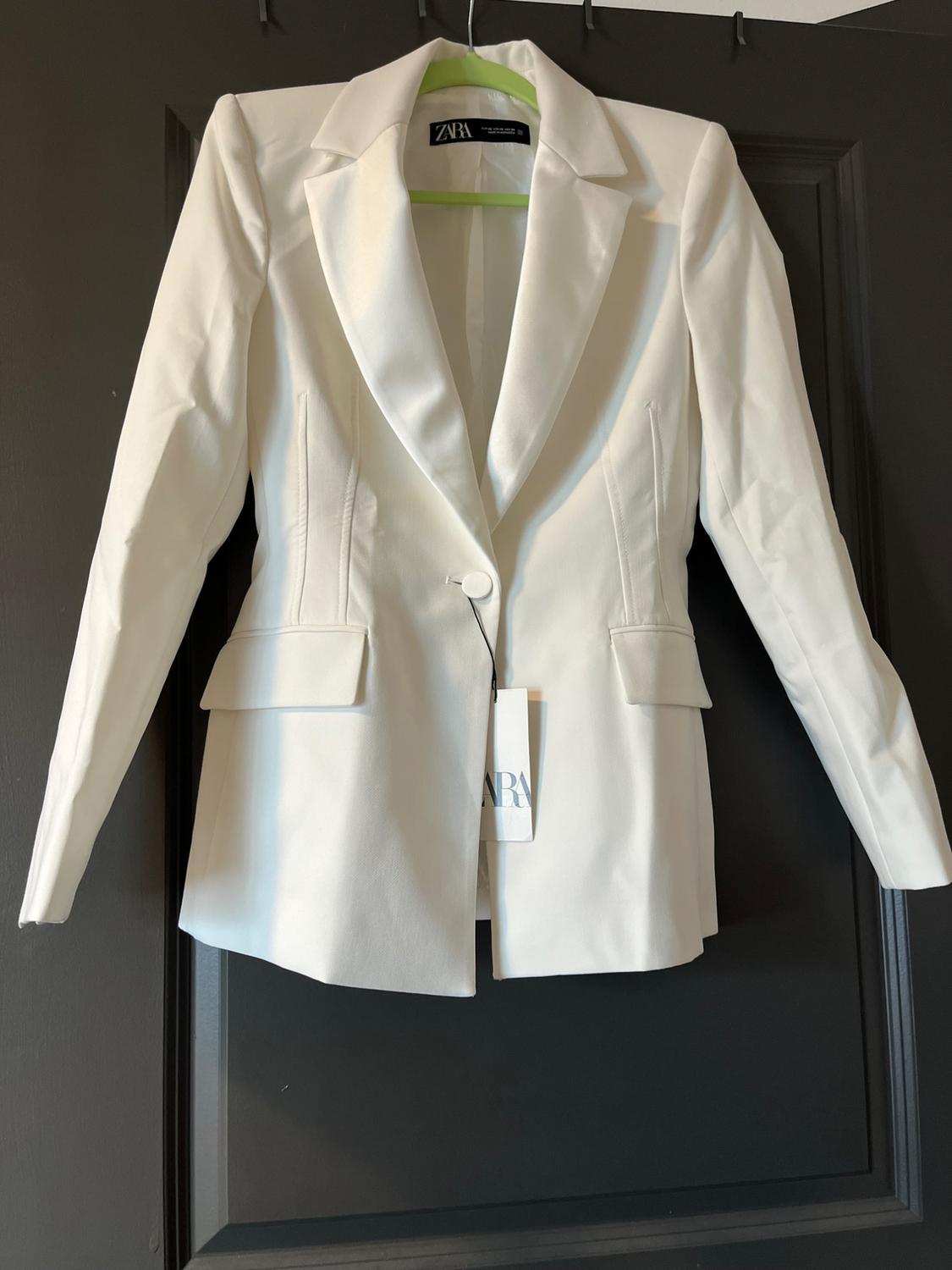 Fitted white blazer with one button and pockets, hanging on a green hanger over a dark door. Visible text: ZARA.