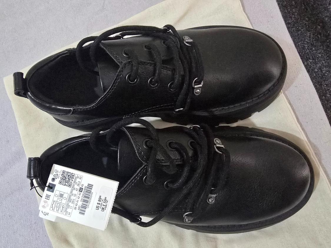 Black lace-up shoes on light-coloured fabric. Label with text: US $ 89.90 UK £ 59.99 ART: 2412/620/800.
