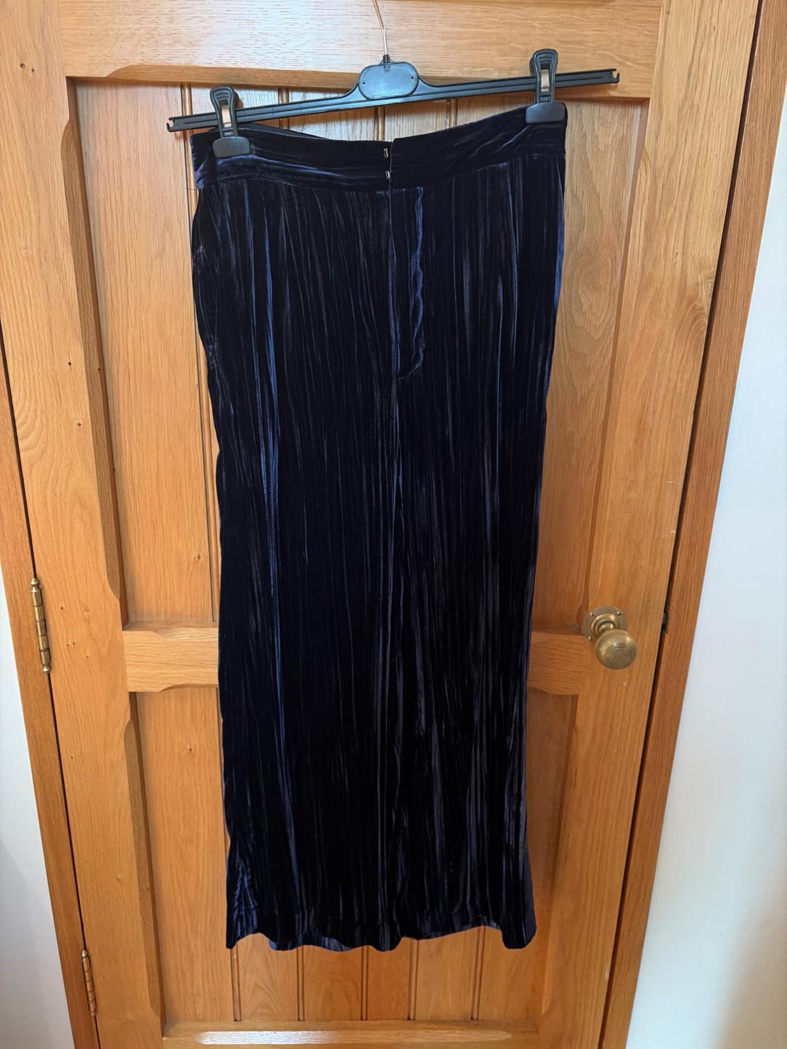 Dark blue pleated velvet maxi skirt hanging on a black hanger over a light wooden door.