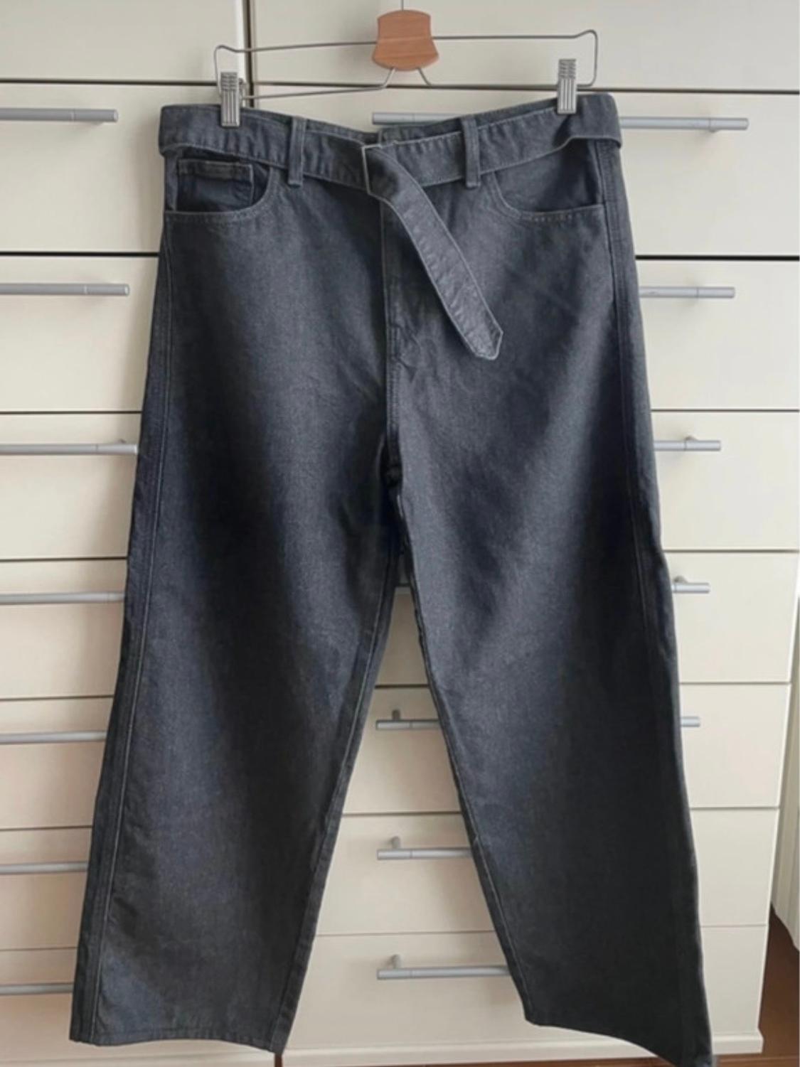 Grey jeans hanging on a hanger in front of a background of white drawers. Matching belt included.