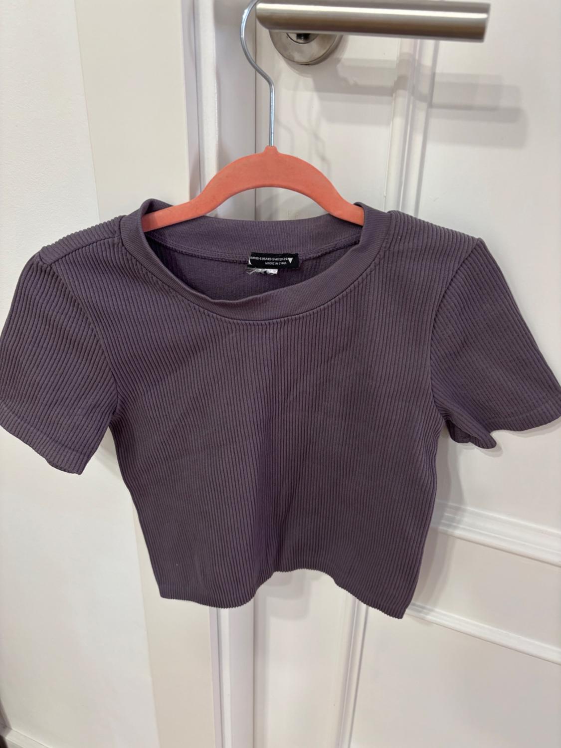 Dark lilac ribbed knit T-shirt hanging on a pink hanger over a white door. Label visible at the neck.
