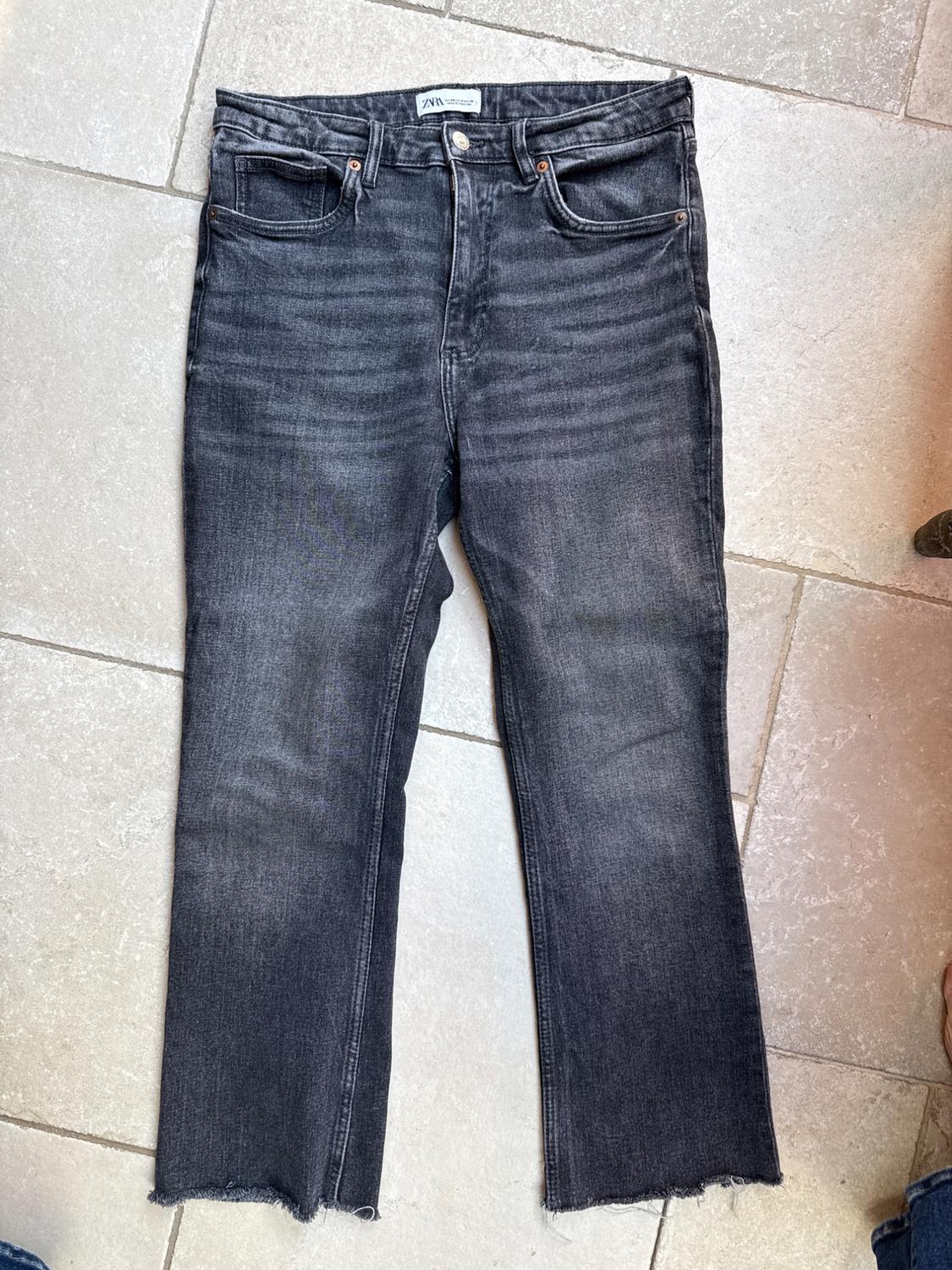 Dark grey distressed jeans with a frayed hem, laid out on a light tiled floor. ZARA label.