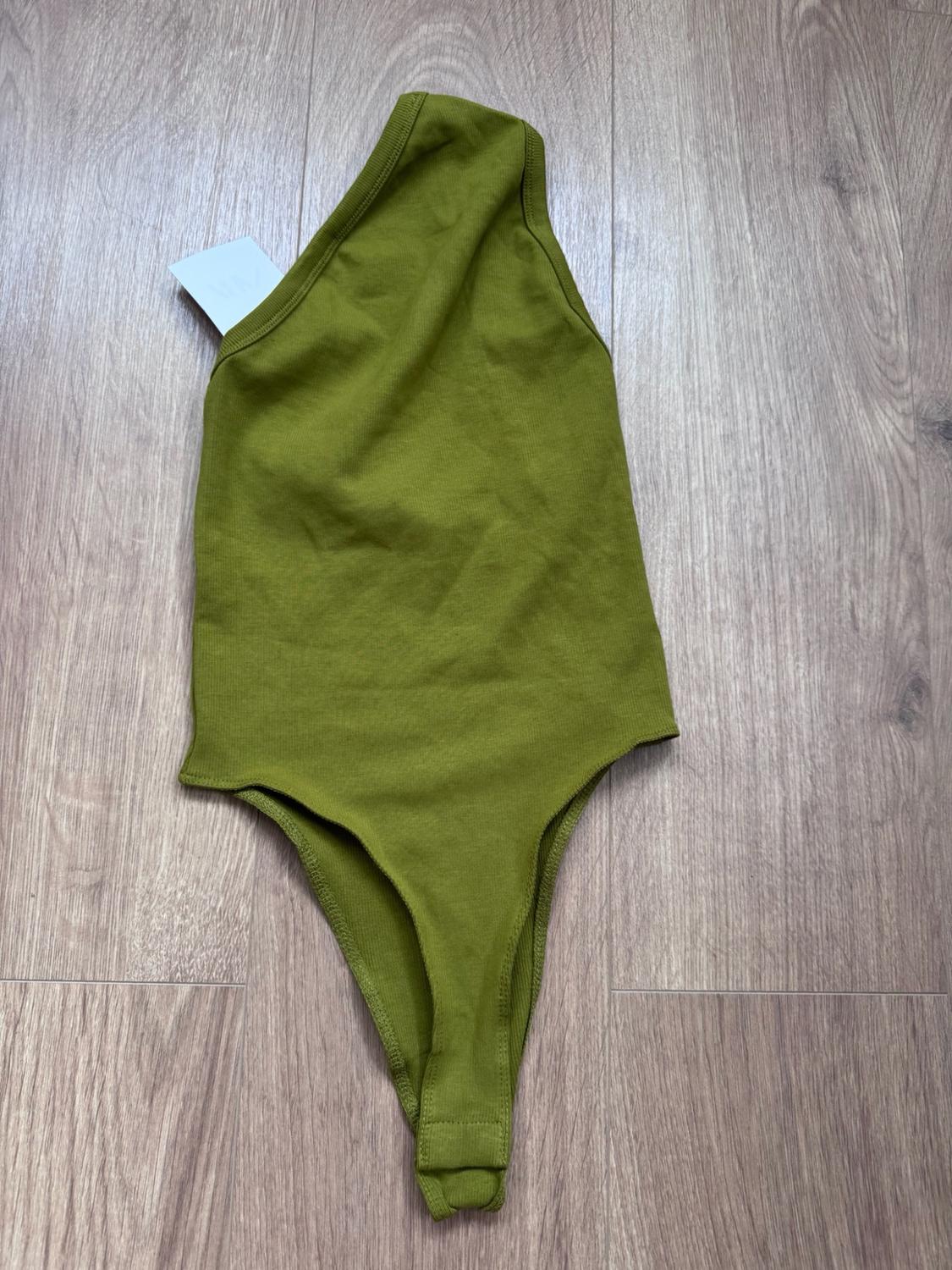 Olive-green asymmetric one-shoulder bodysuit with a white label, photographed on a light wooden floor.