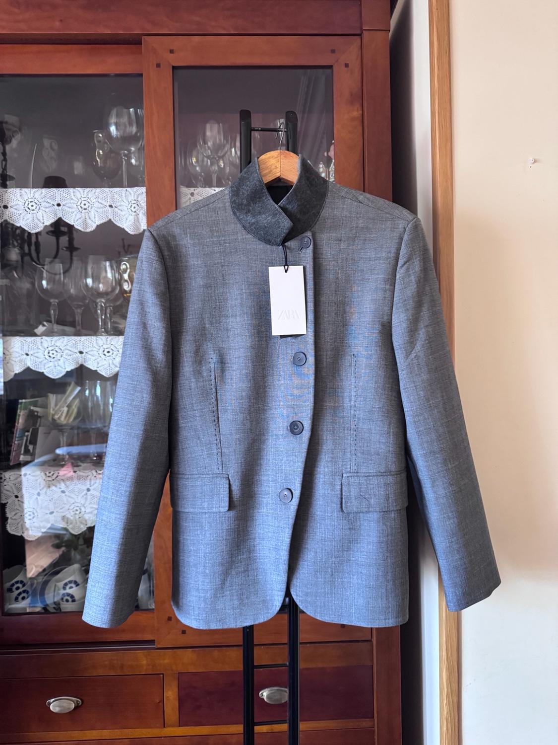 Grey buttoned jacket with a dark collar, hanging on a coat hanger in front of a wooden display cabinet. Label with the text ZARA.