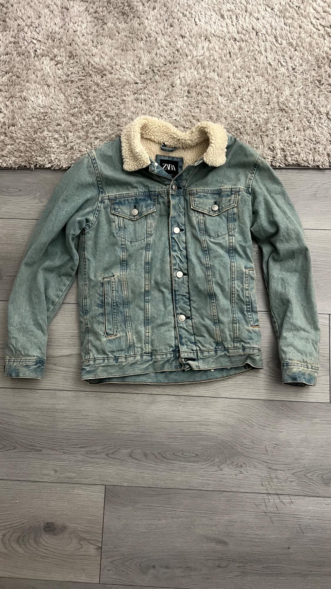 Light blue denim jacket with a white fur collar and a ZARA tag, on a gray wooden floor and a rug.