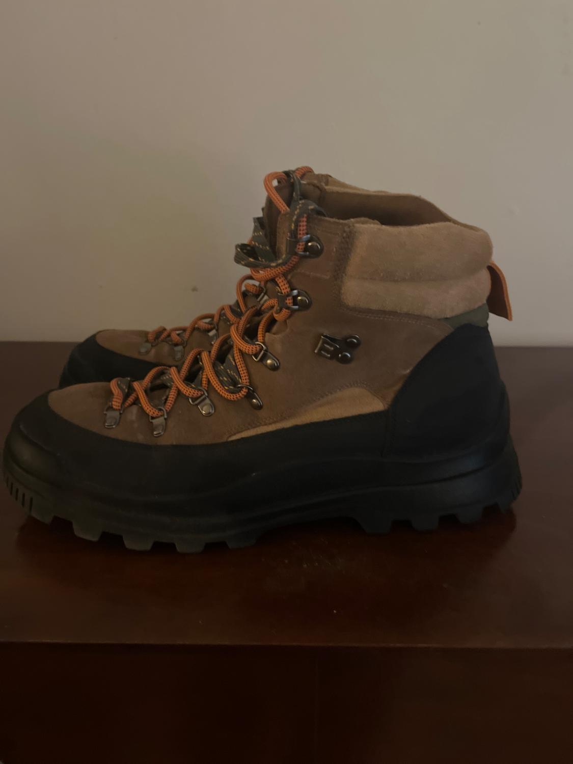 Brown hiking boots with black soles and orange laces on a wooden surface, white background.