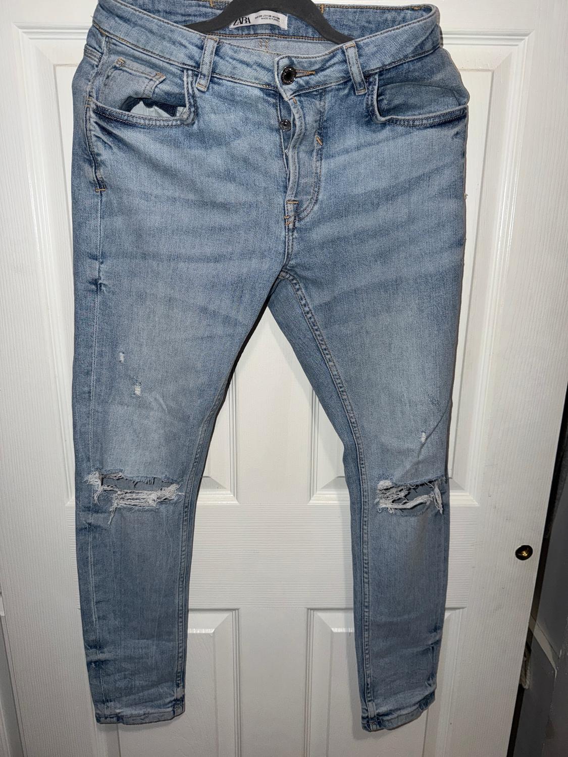 Worn blue jeans hanging on a black hanger in front of a white door. Ripped details on the knees.