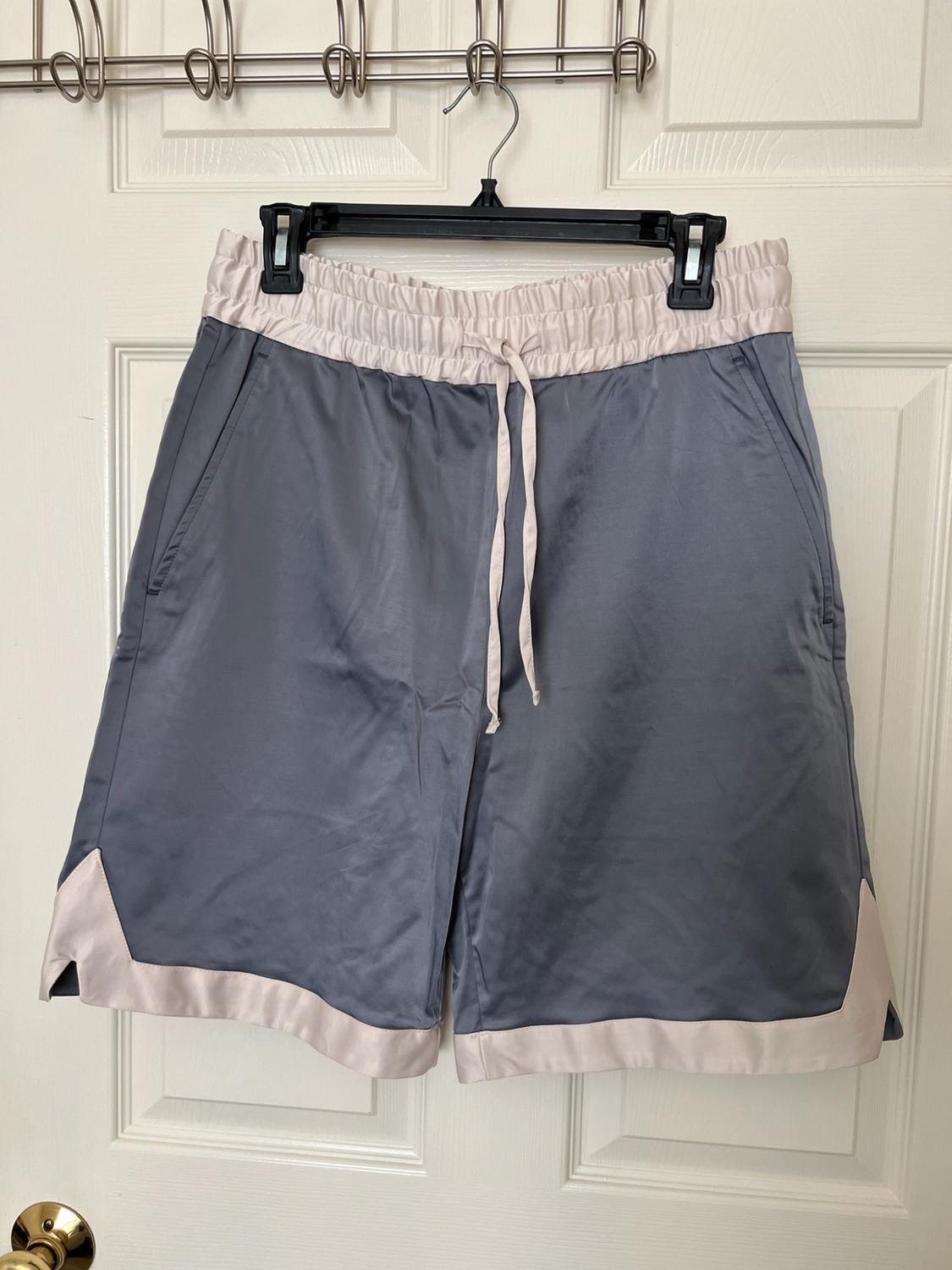 Grey satin shorts with an elasticated waistband, drawstring and beige trim, hanging on a black hanger over a white door.