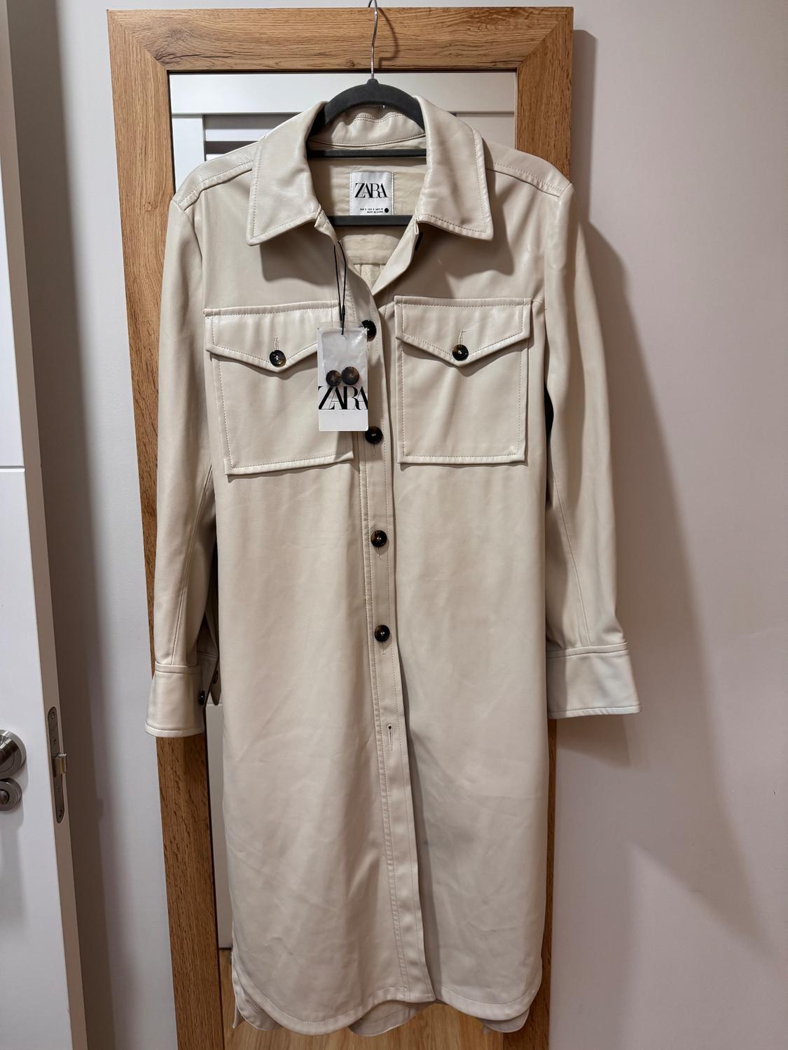 Long-sleeved beige coat with front pockets and black buttons, hanging on a hanger with a Zara tag.