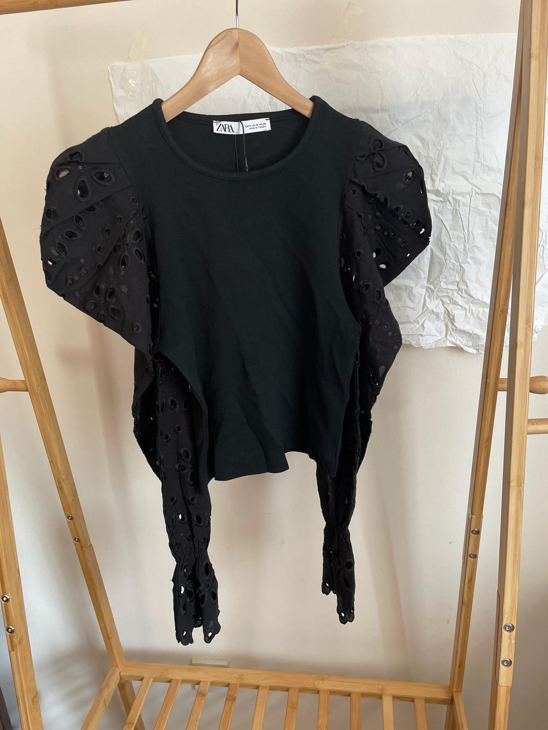 Black ZARA top with long puffed sleeves and eyelet embroidery, hanging on a wooden clothes rail. Crinkled white paper background.