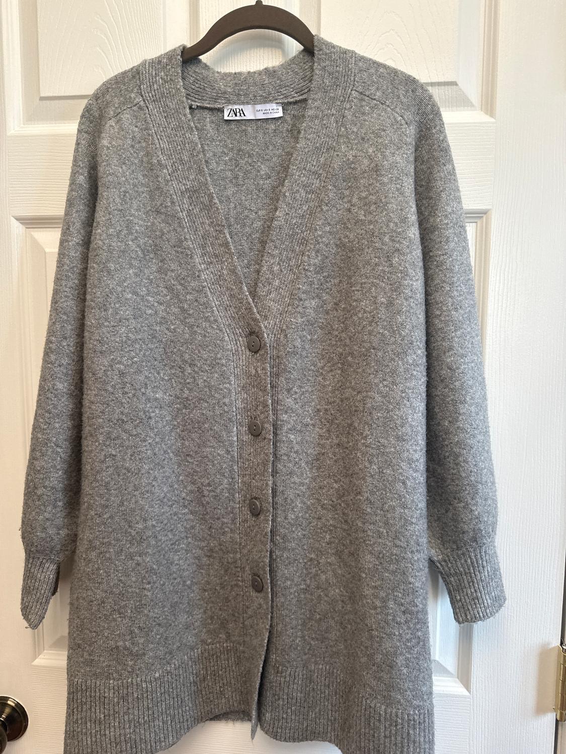 Grey knitted cardigan hanging on a hanger in front of a white door. Visible label with ZARA brand.
