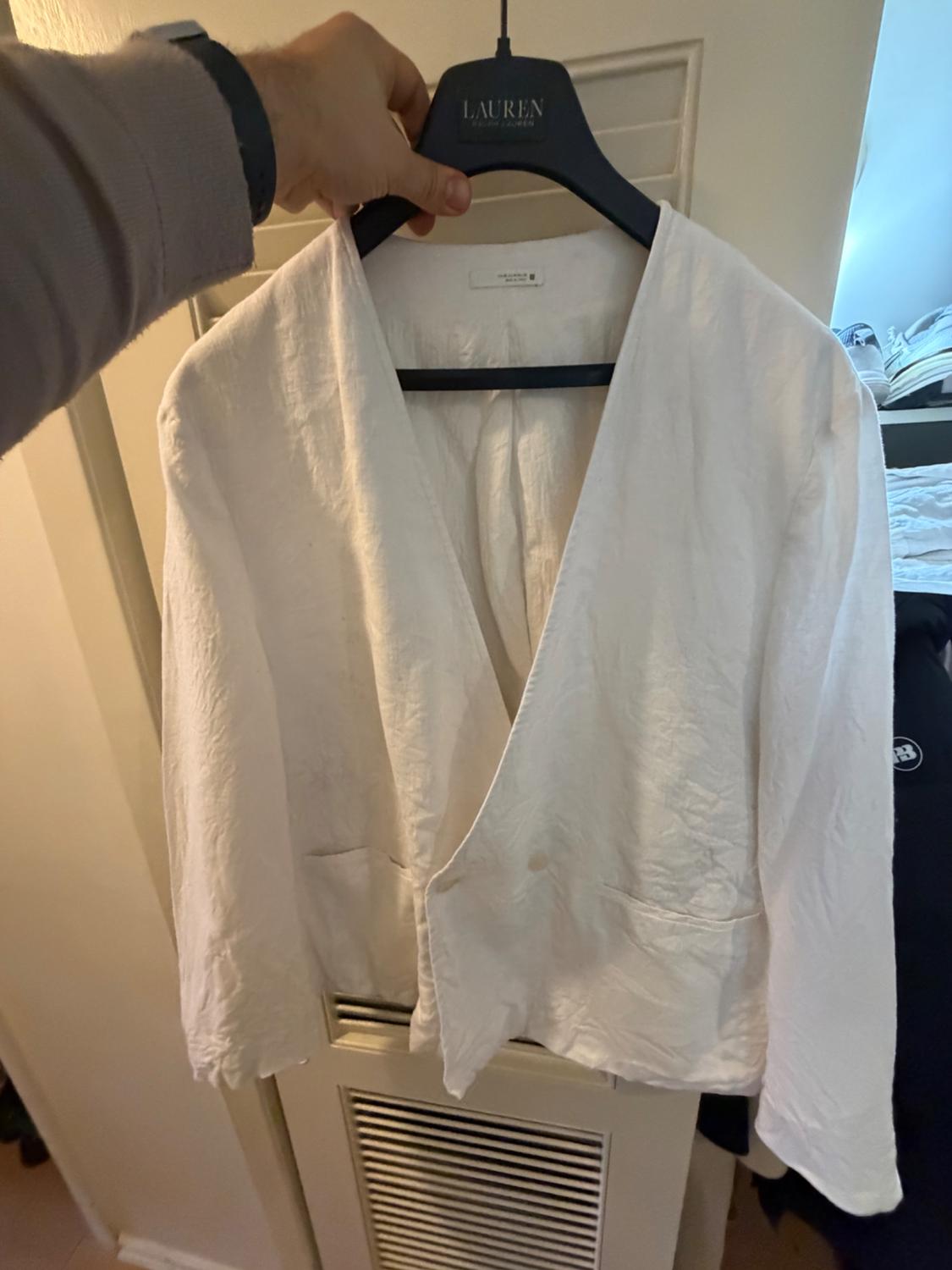 White linen jacket hanging on a black hanger with the brand visible. Hand holding the hanger in an indoor setting.