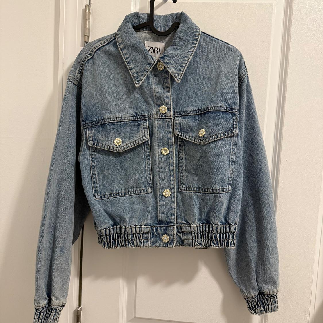 Blue denim jacket with decorative flower-shaped buttons, front pockets, and elastic cuffs and waistband, hanging on a hanger.