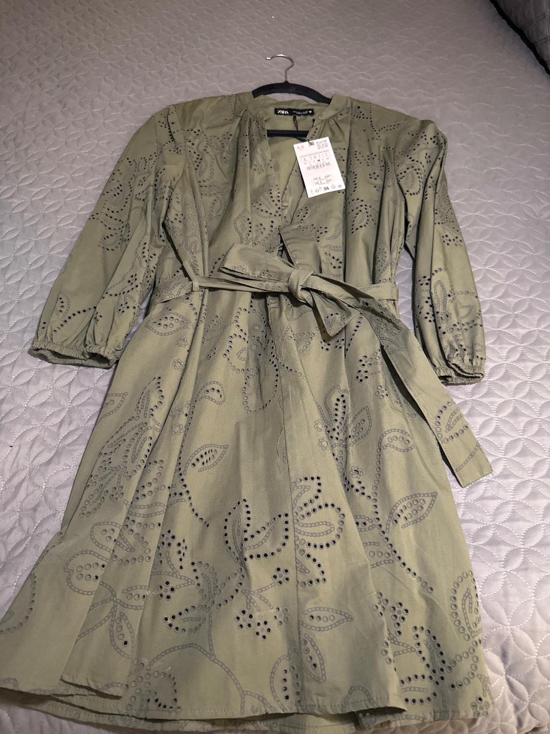 Olive green dress with perforated floral print, long sleeves, and matching belt, hanging over a gray quilted comforter.