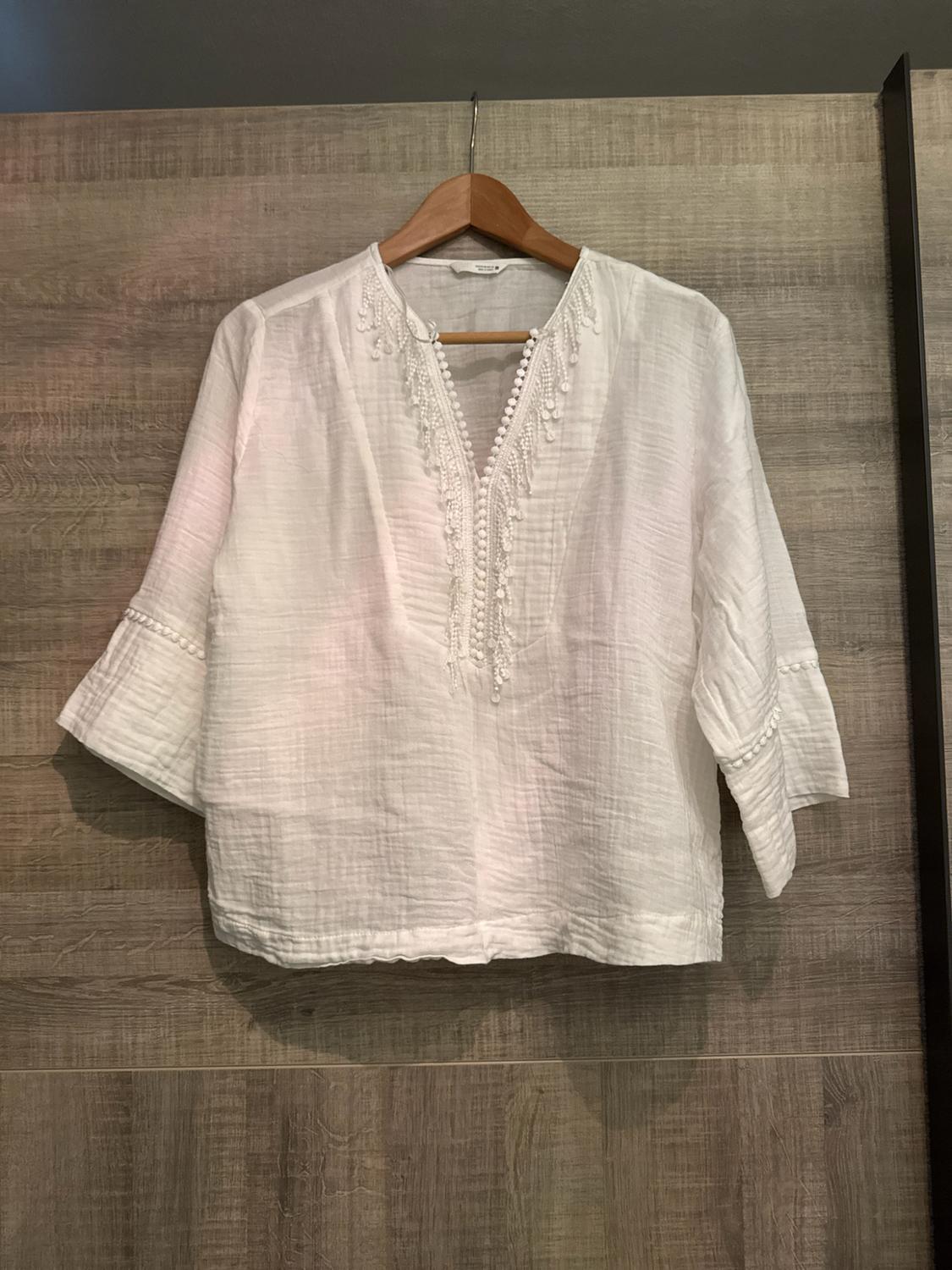 White blouse with a V-neck and fringe detailing on the neckline and sleeves, hanging on a hanger against a wooden background. Relaxed style.