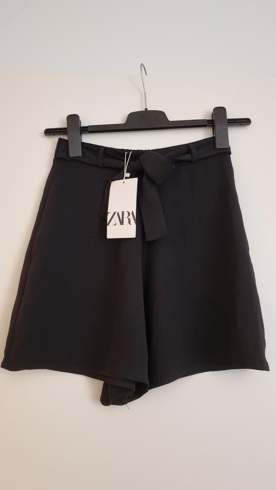 Black shorts hanging on a hanger with a belt and Zara tag. White background.