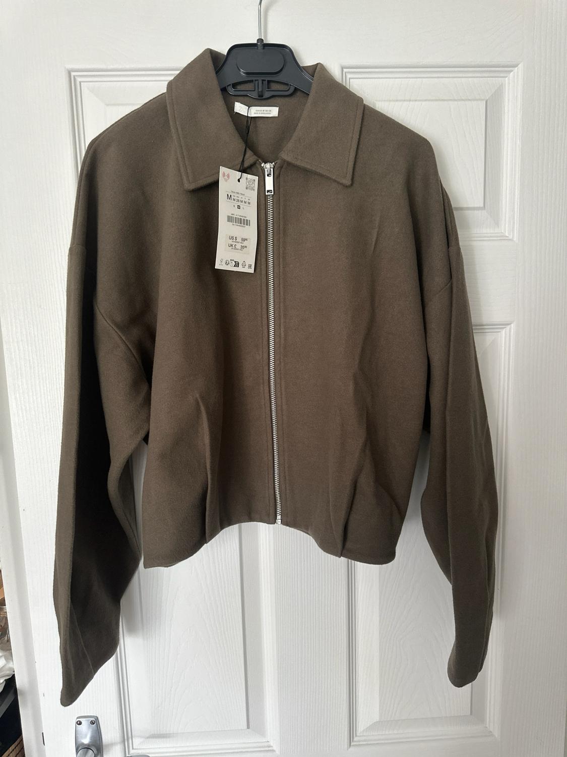 Brown jacket with a silver zip and collar, hanging on a black hanger over a white door.
