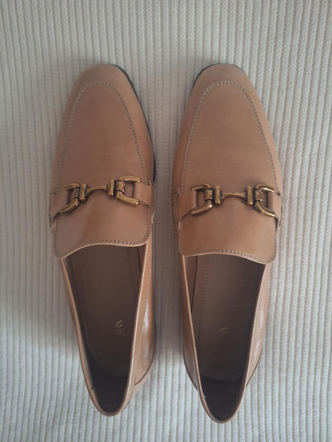 Light brown leather shoes with a gold metallic detail on the upper, on a textured beige fabric background.