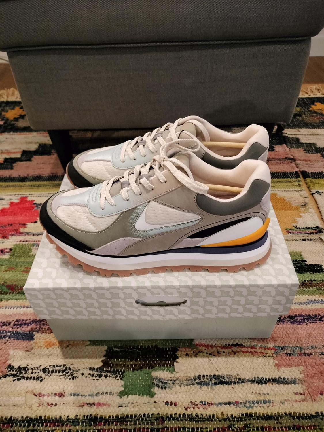 Grey and white trainers on a box with a geometric pattern, placed on a colourful rug.