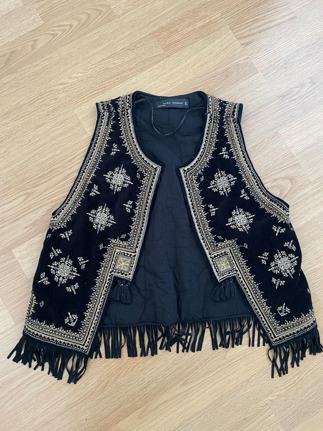 Black waistcoat with gold embroidery and fringing on the trim, on a light wooden floor. Visible label: "ZARA WOMAN".