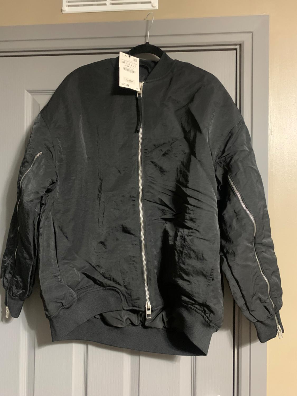 Black jacket hanging on a grey door, with a front zip and a visible white label on the collar. Indoor setting.