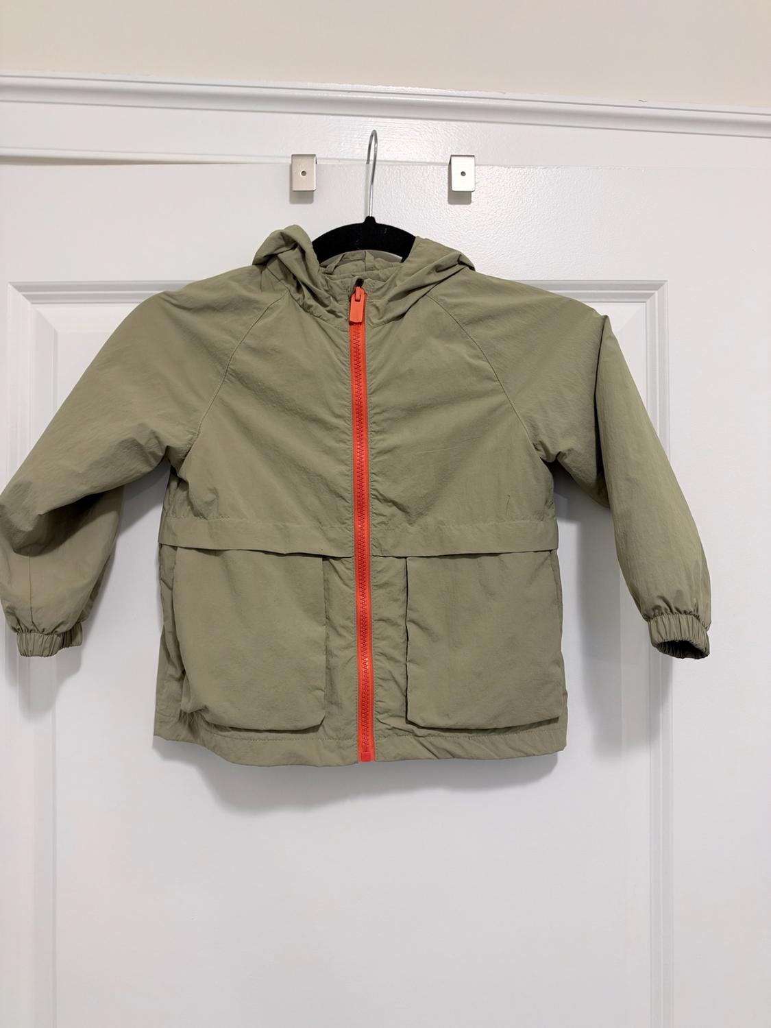 Olive green hooded jacket, with orange front zipper and pockets, hanging on a hanger on a white door.