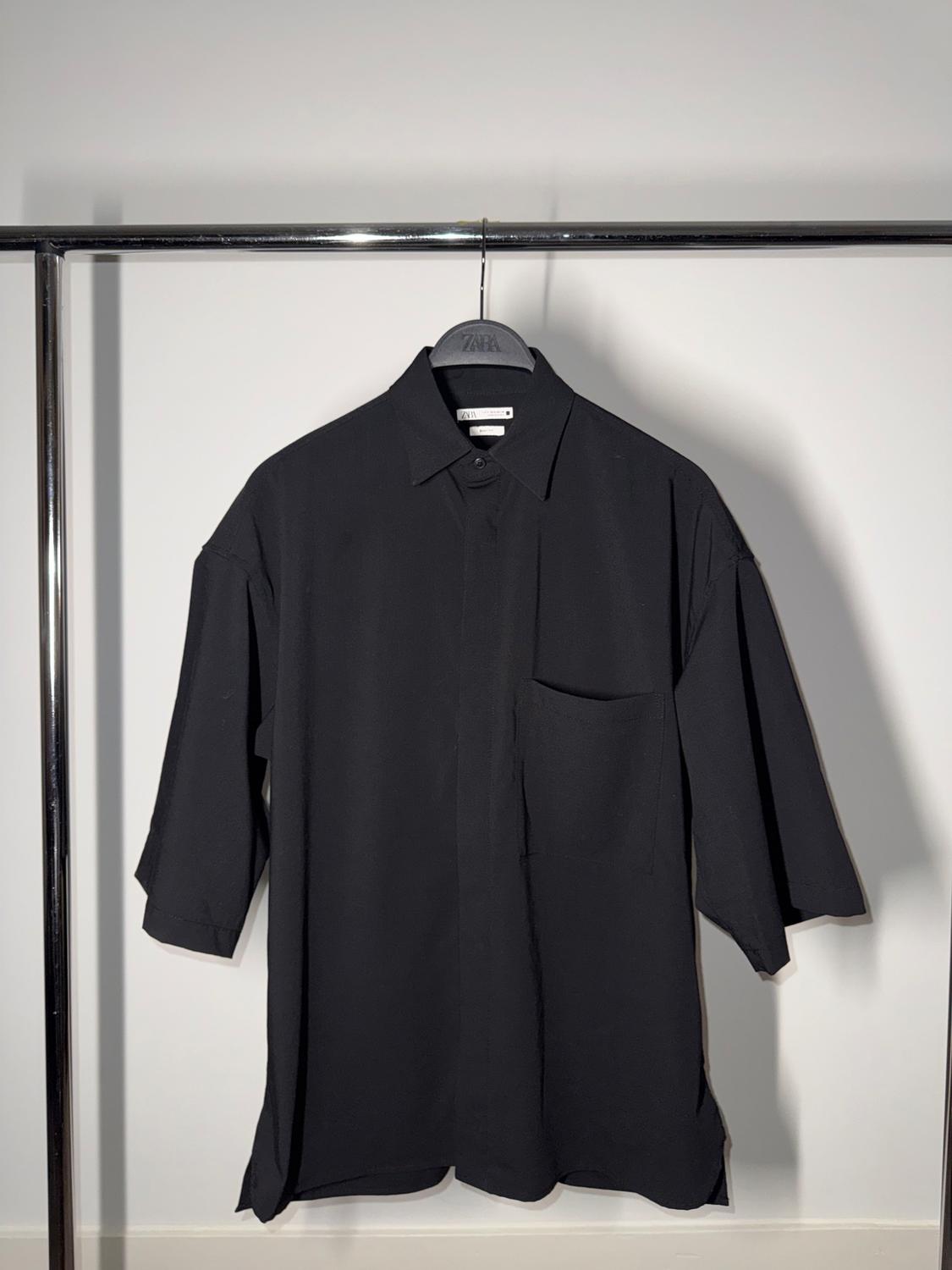 Black short-sleeved shirt hanging on a metal clothes rail, with a chest pocket and classic collar. White background.