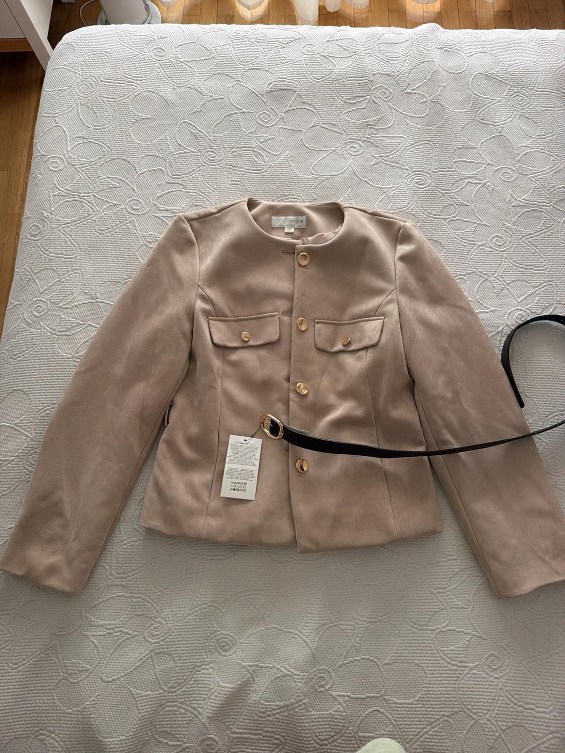 Beige jacket with gold buttons and pockets, alongside a black belt, on a bed with a white floral bedspread. Relaxed style.