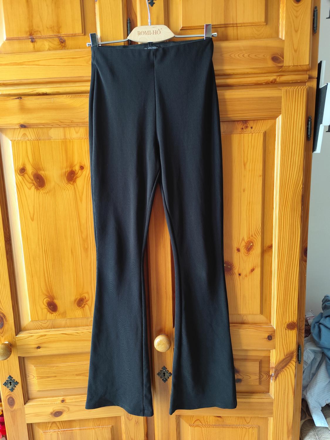 Black flared trousers hanging on a "ROMI-HO" branded hanger, over a light wooden door.