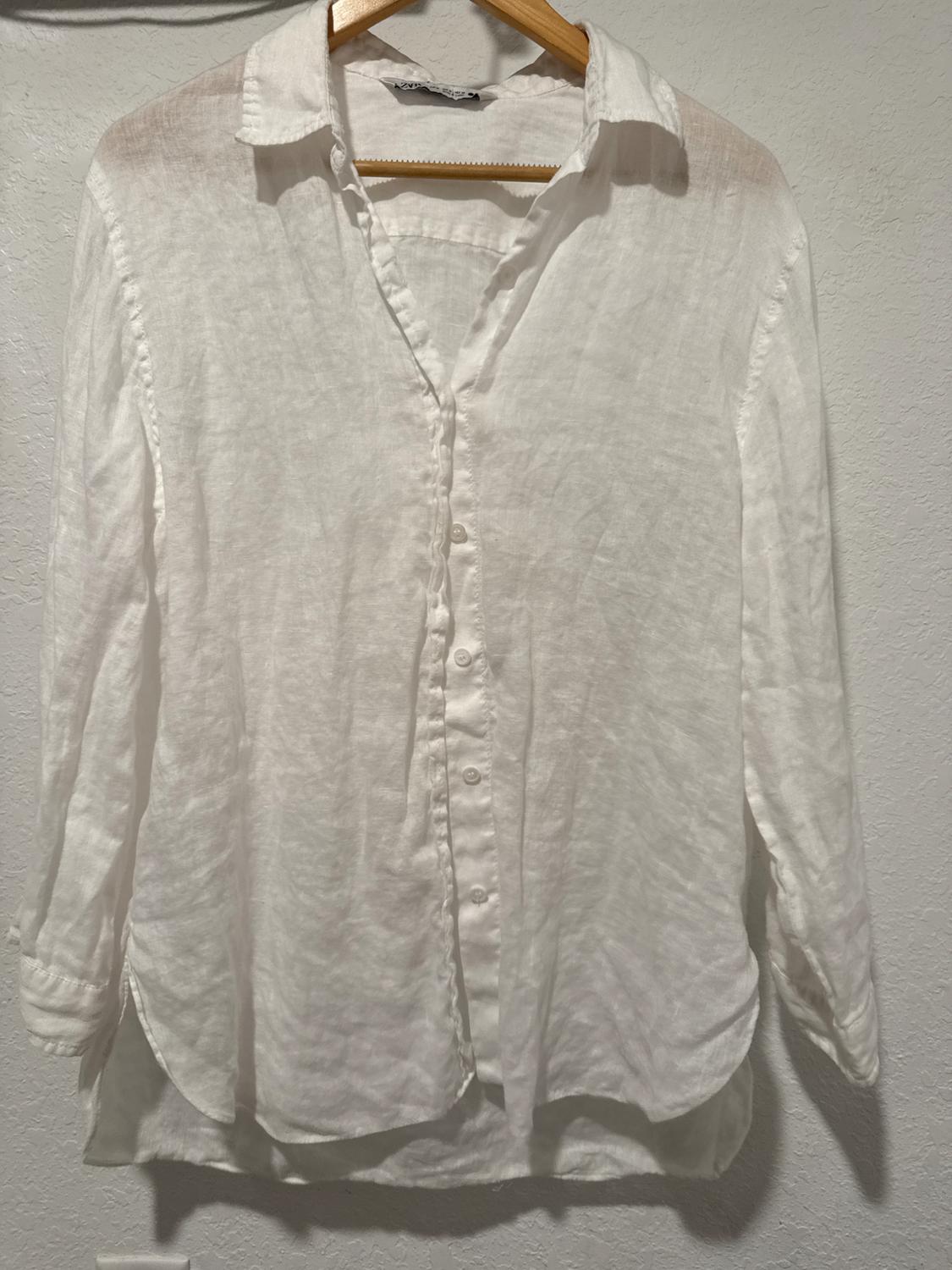 White linen shirt hanging on a wooden hanger against a white wall. Long sleeves and buttons at the front.
