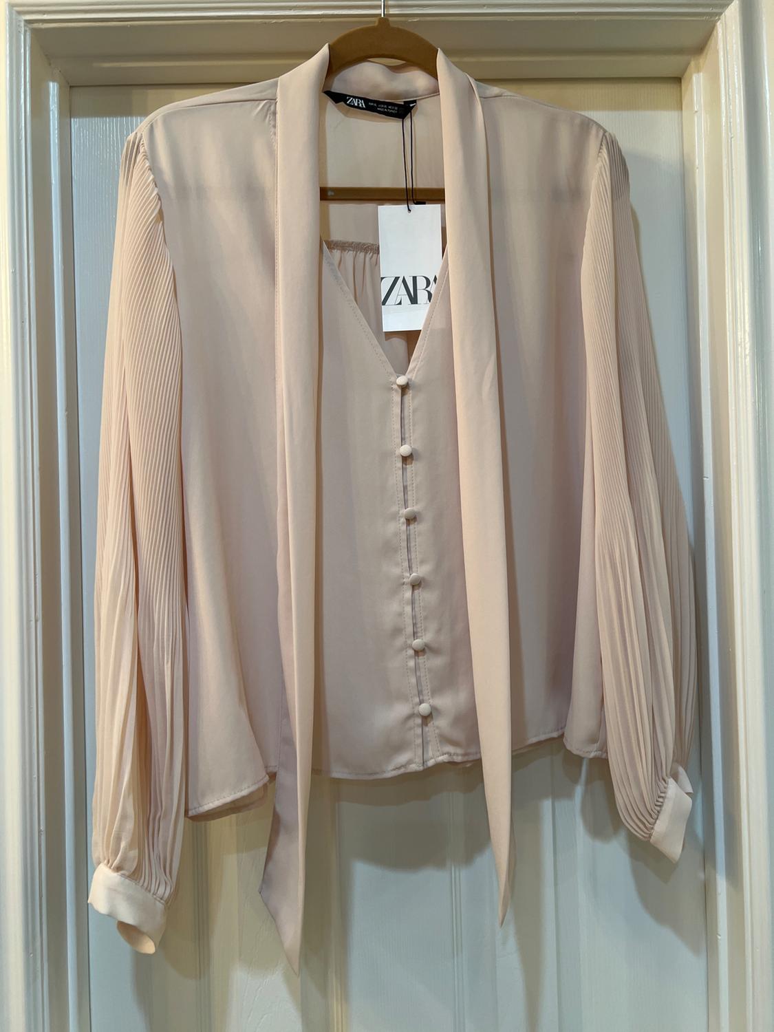 Beige long-sleeved blouse with front buttons and a neck tie, hanging on a wooden hanger. Visible label: ZARA.