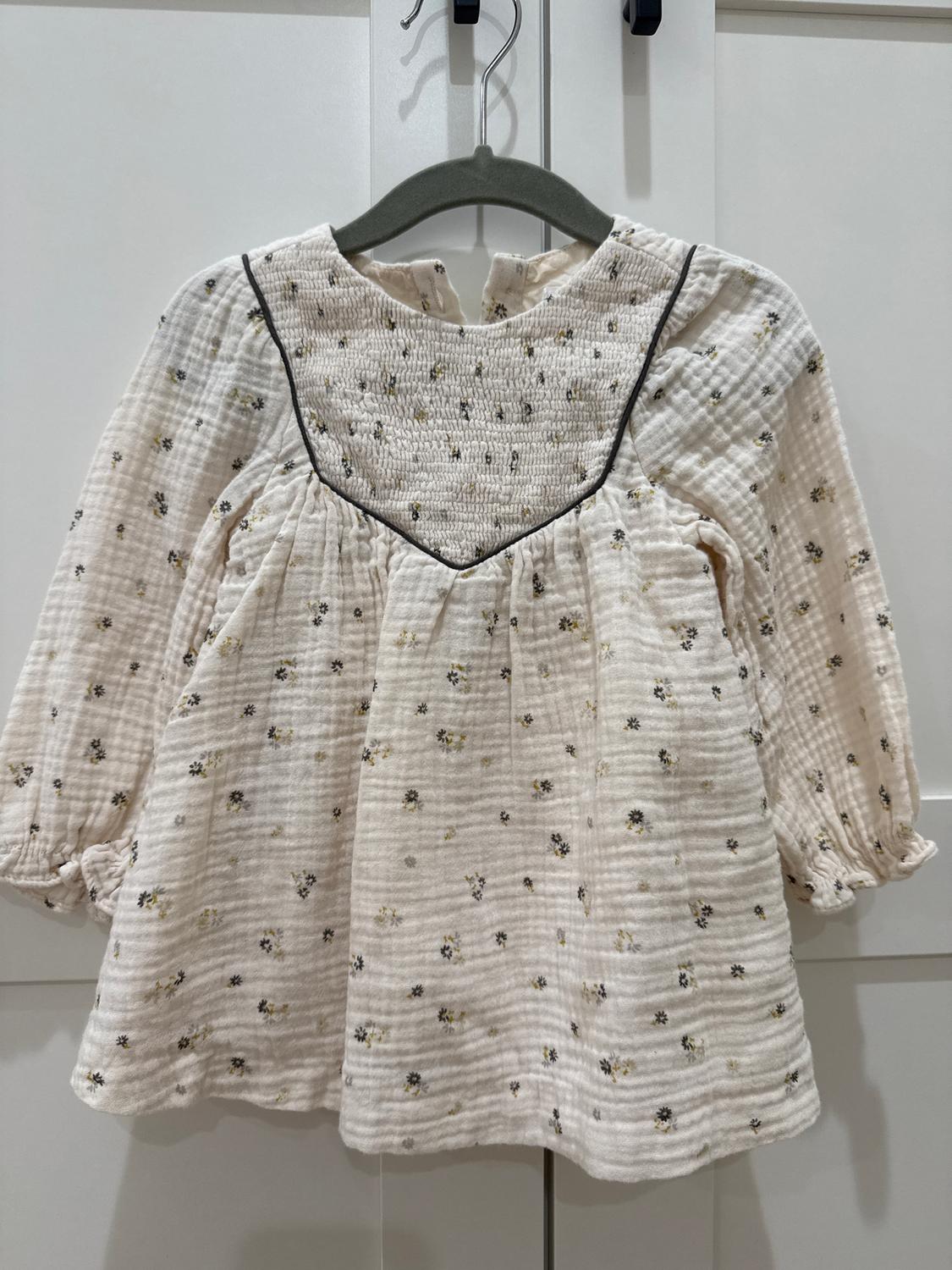 White children's dress with a small black and yellow floral print, long sleeves, and dark trim detail, hanging on a hanger.