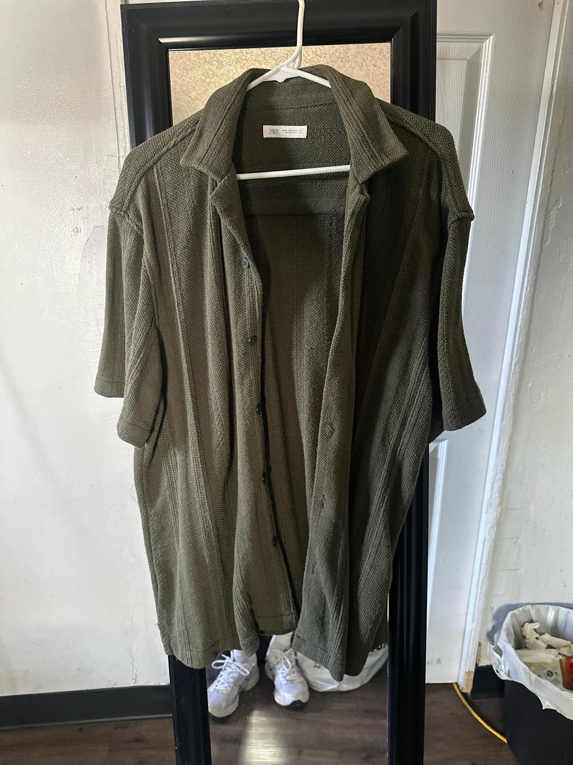Textured olive green short-sleeved shirt, open and hanging on a hanger in front of a mirror. The label reads ZARA.