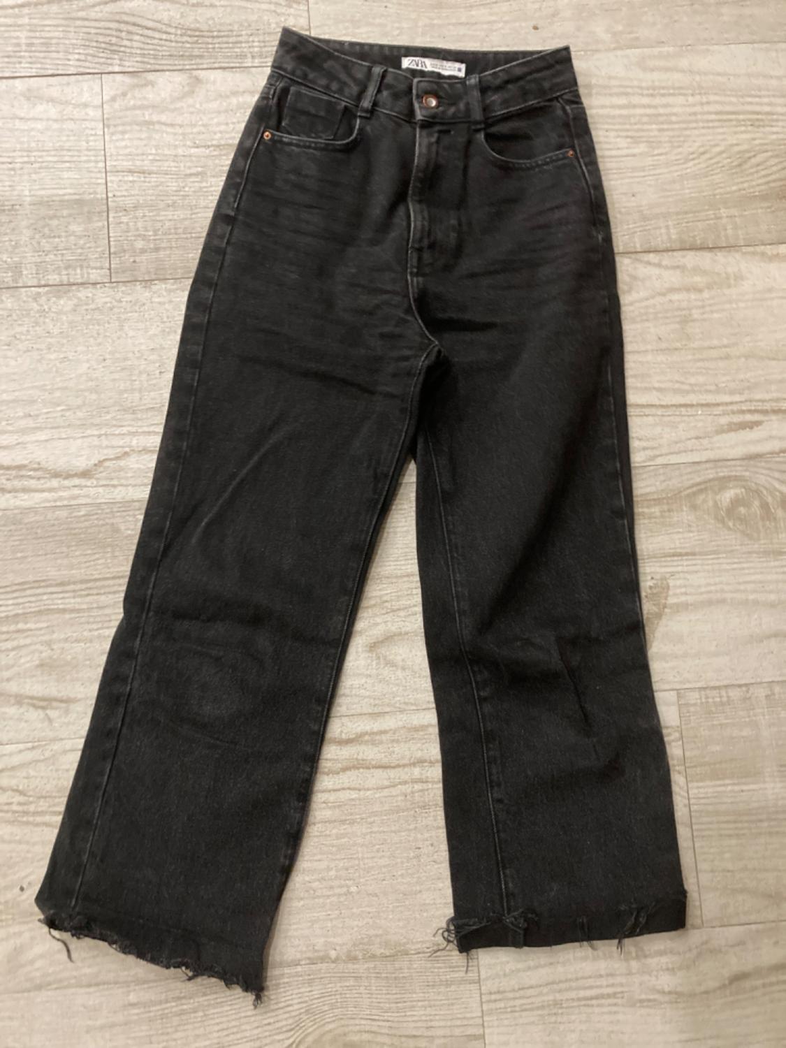Black, straight-cut jeans with a frayed hem, laid out on a light wooden floor.