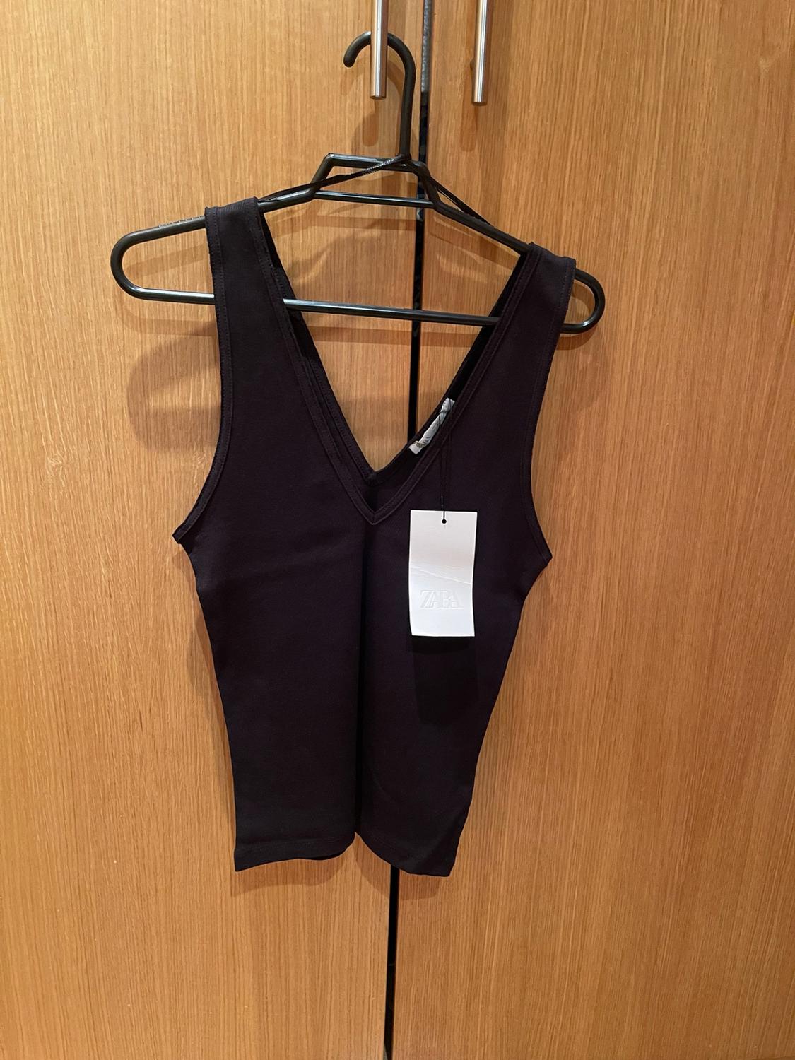 Black sleeveless V-neck top, hanging on a hanger against a wooden background. Includes a white label with the text ZARA.