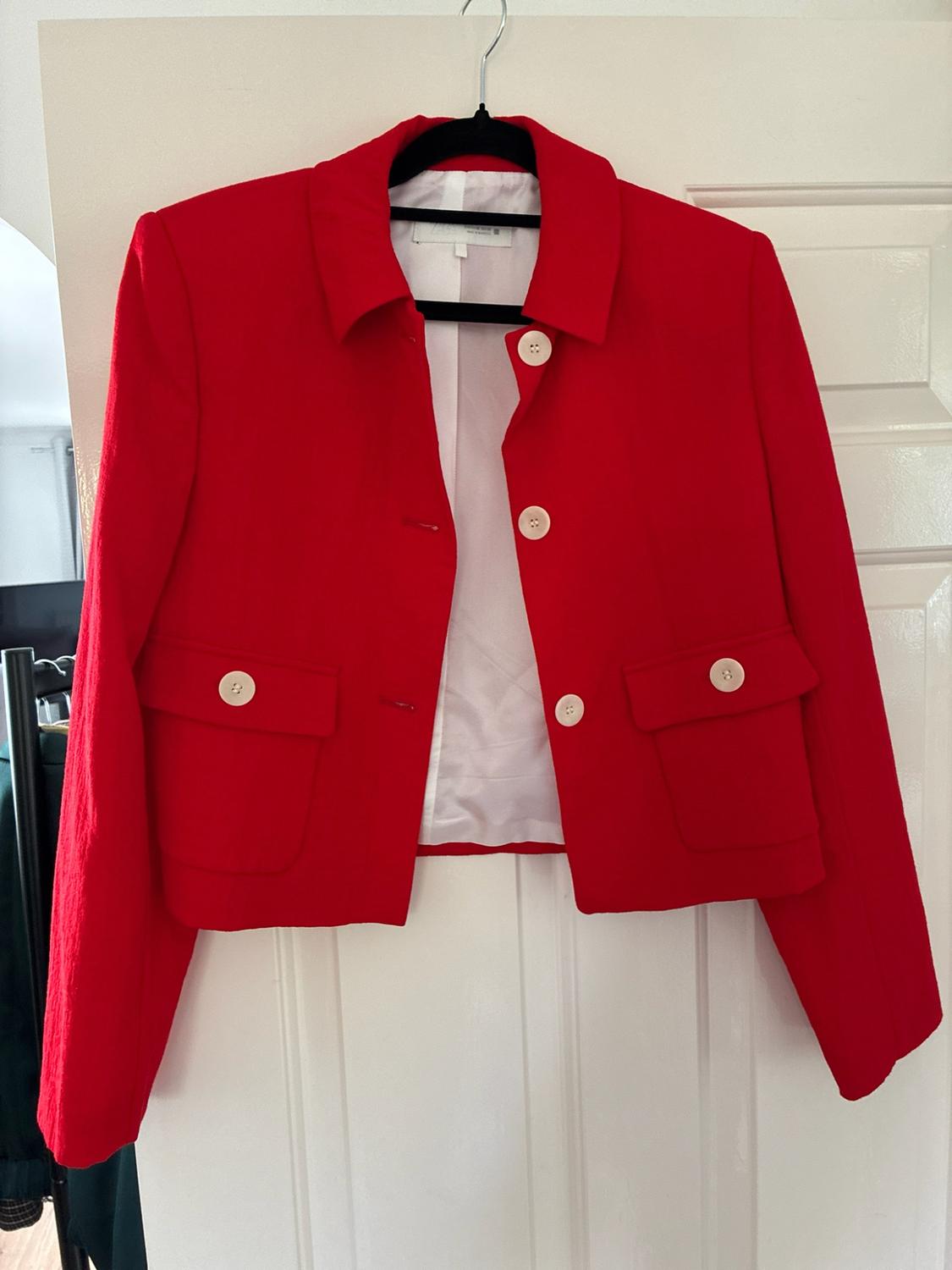 Open red jacket with lining and white buttons, plus front pockets, hanging on a hanger on a white door.