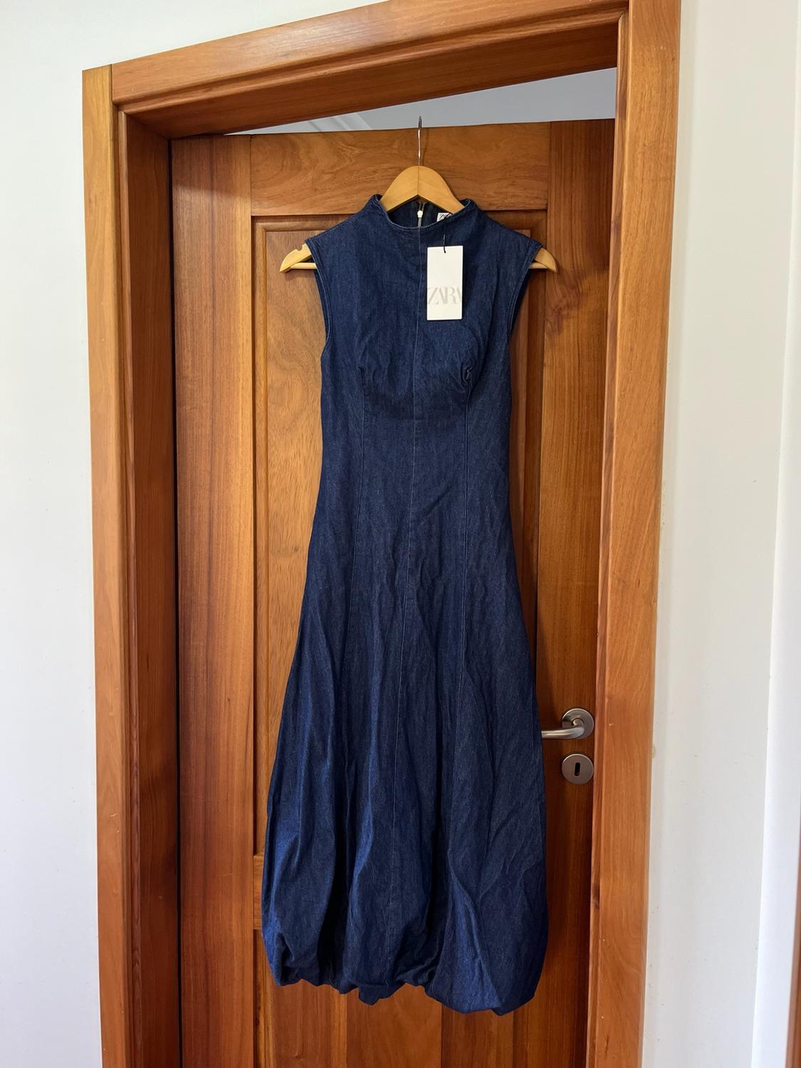 Dark blue denim dress, high neck, sleeveless with a puffed hem, hanging on a wooden door. Label with the text ZARA.