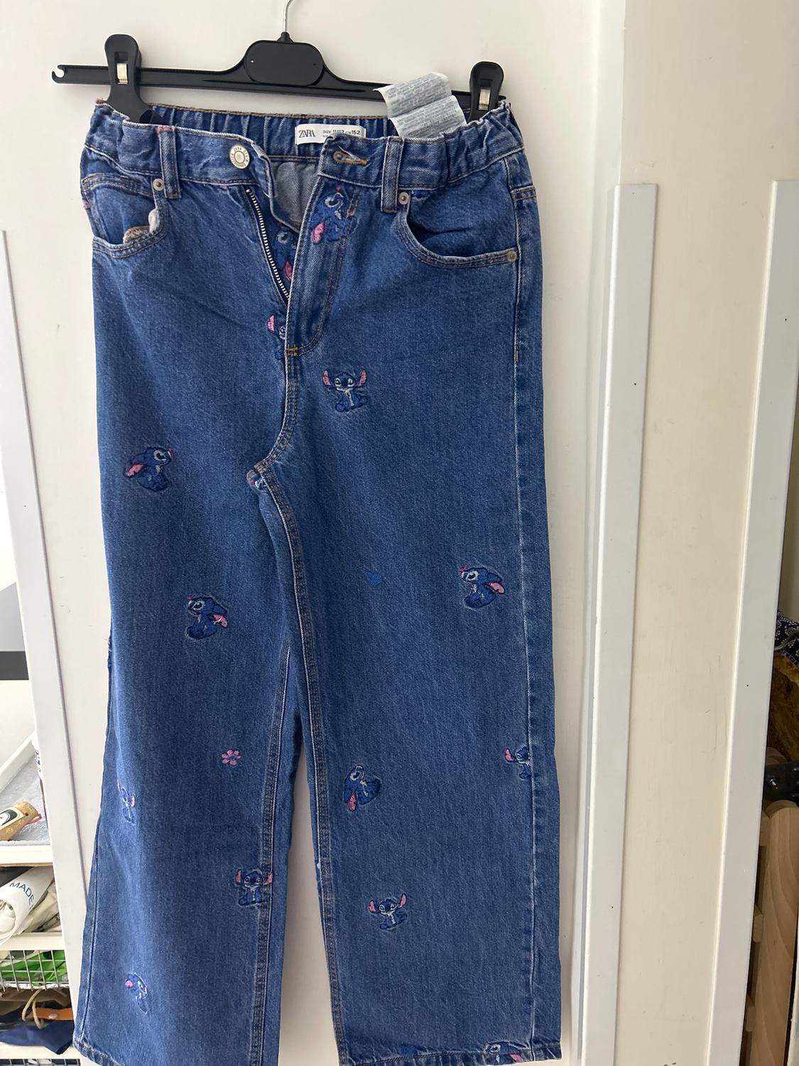 Blue denim jeans with pink and blue embroidered cartoon character, hanging on a black hanger.
