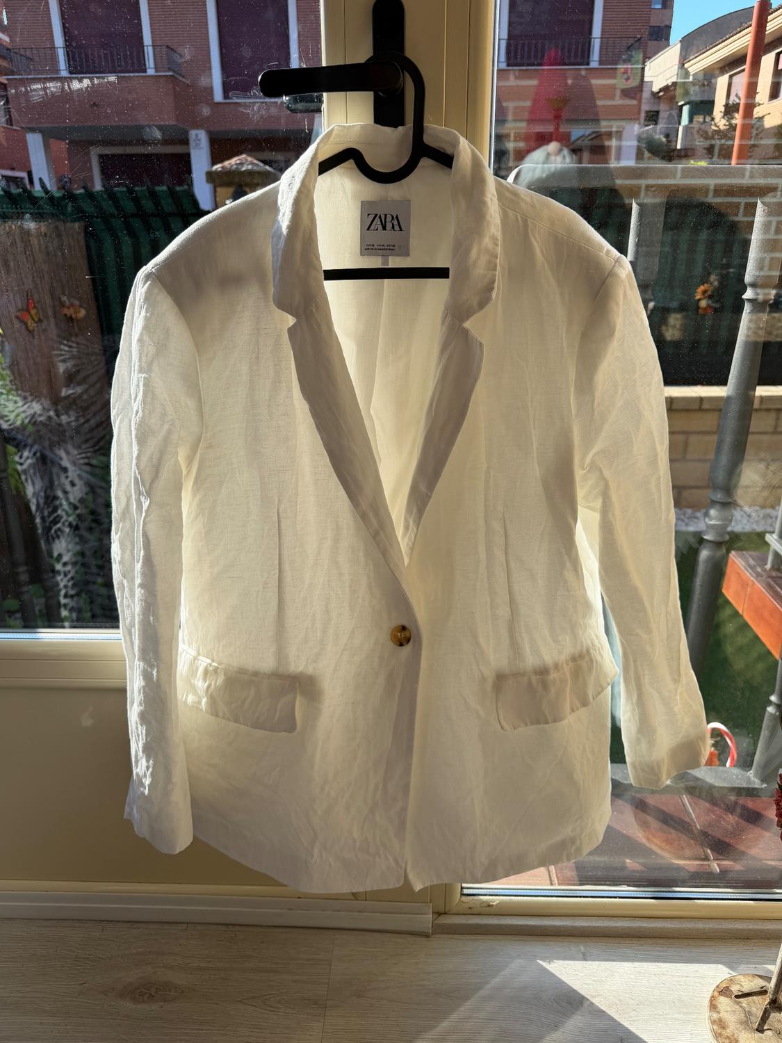 White jacket hanging on a hanger in front of a window, with a visible Zara tag. Background of buildings and an outdoor garden.