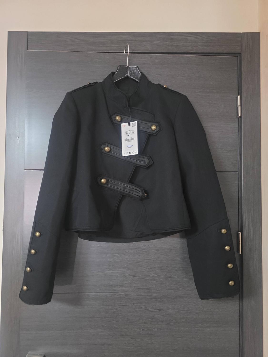 Short black military-style jacket with gold buttons and a high collar, hanging on a hanger over a grey door.