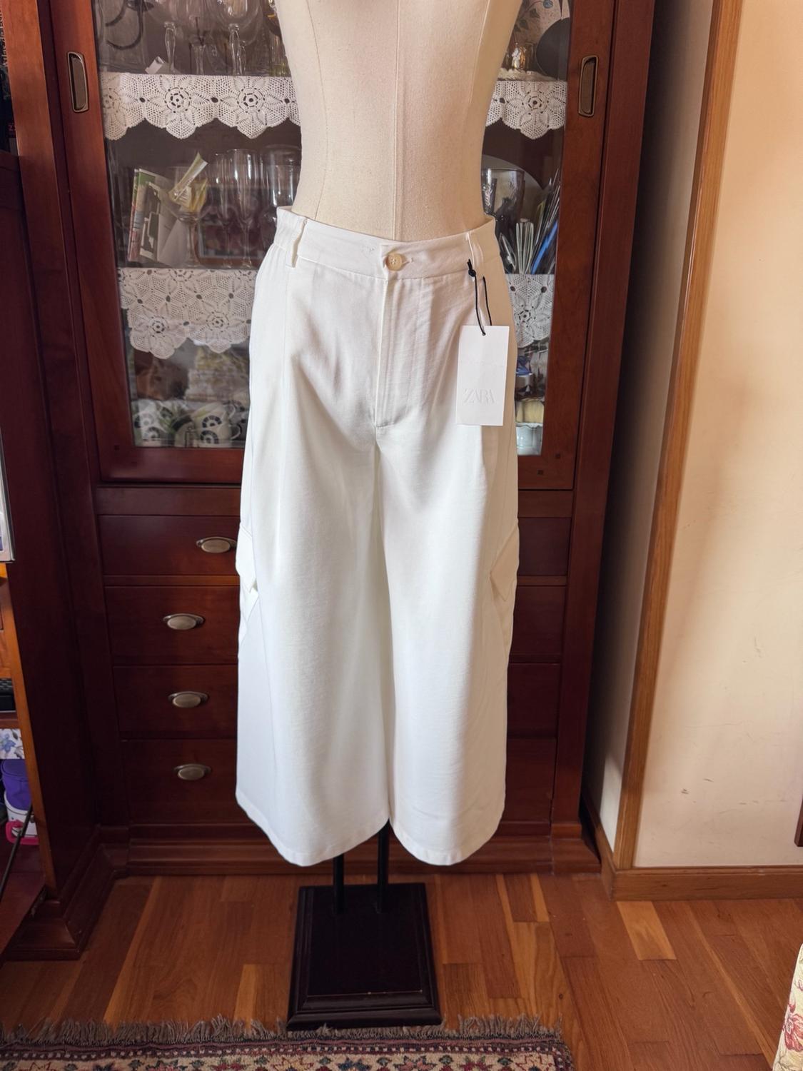 Mannequin wearing wide-leg cropped white trousers with pockets and a ZARA label. Interior background with a classic wooden display cabinet and parquet flooring.
