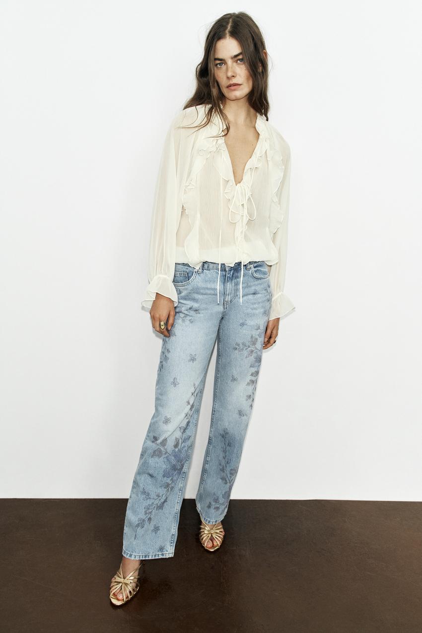White blouse set with ruffles and a tie neckline, and light blue jeans with a floral print on the legs.