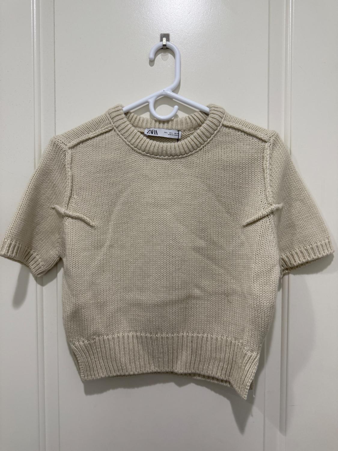Beige short-sleeved sweater hanging on a white hanger over a white door. Visible label with the ZARA brand.