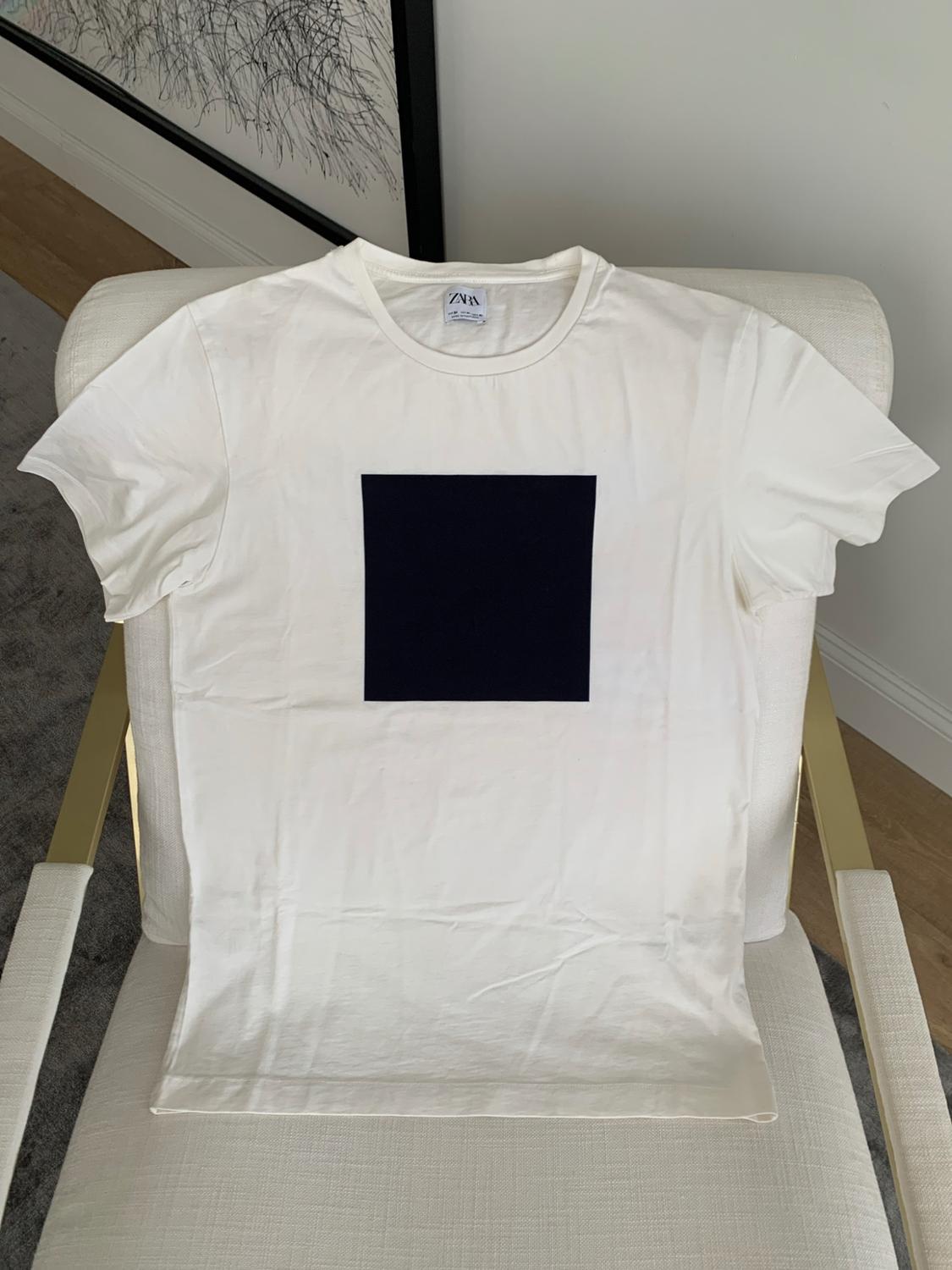 White short-sleeved T-shirt with a black square, on a light-coloured armchair with gold-coloured arms. ZARA label visible at the neck.