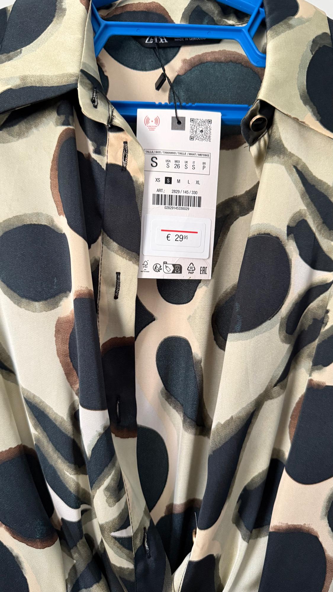 Shirt with a large circle print in dark and light tones, hanging on a blue hanger. Label shows price of €29.95.
